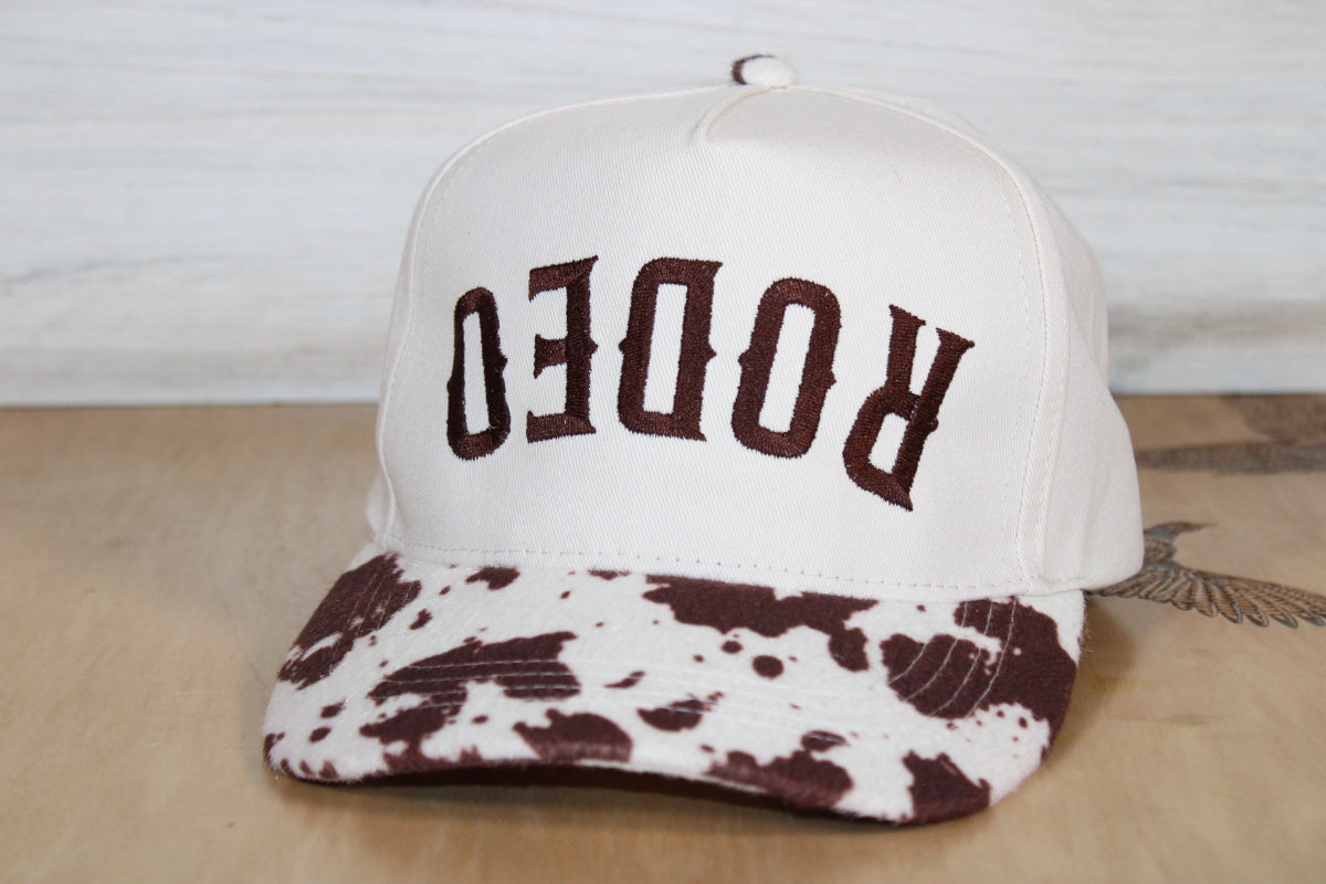 White cap with brown cow print brim and 'RODEO' text on a wooden surface.