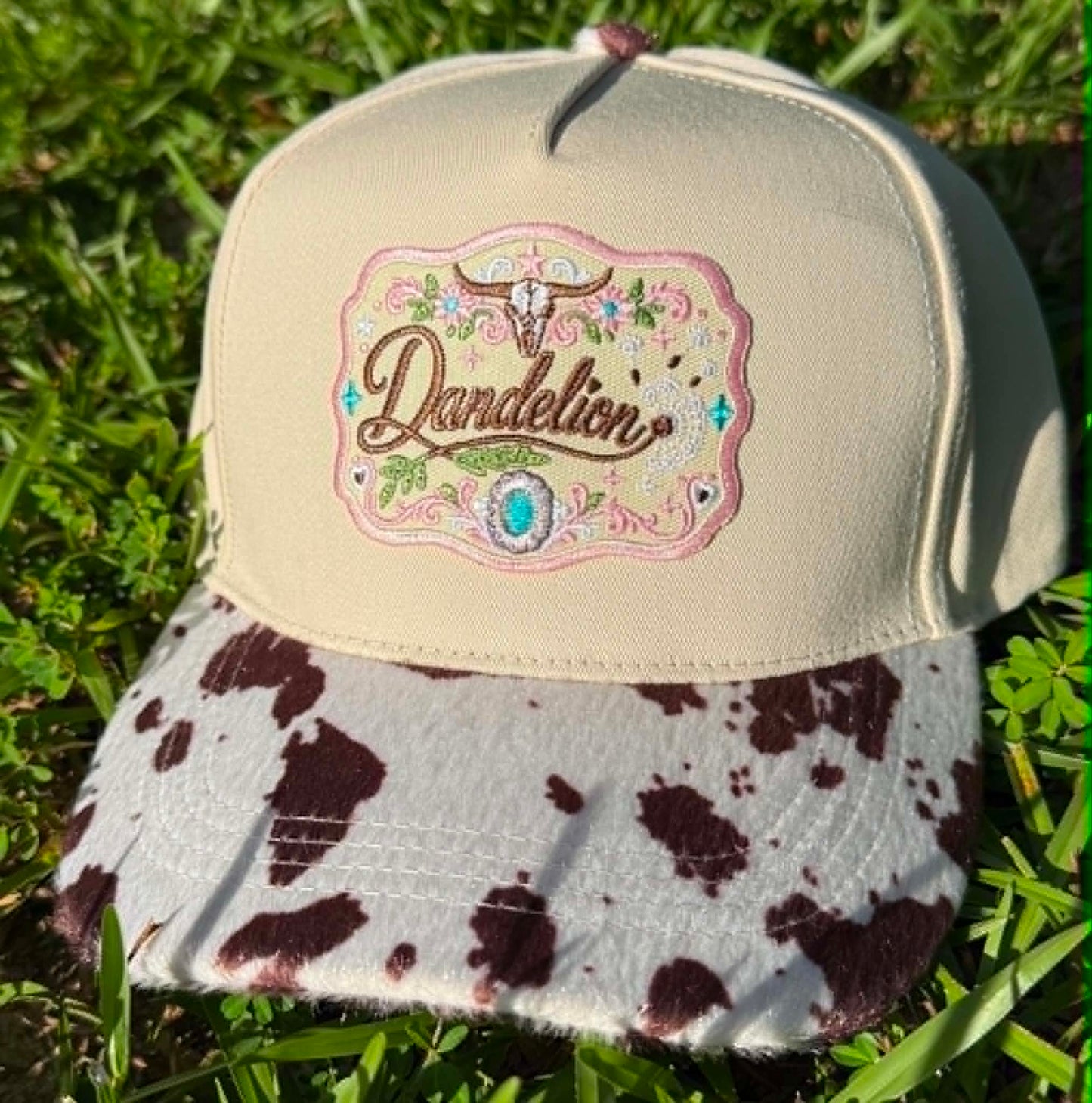 Beige cap with cow print brim and 'Dandelion' logo on grass