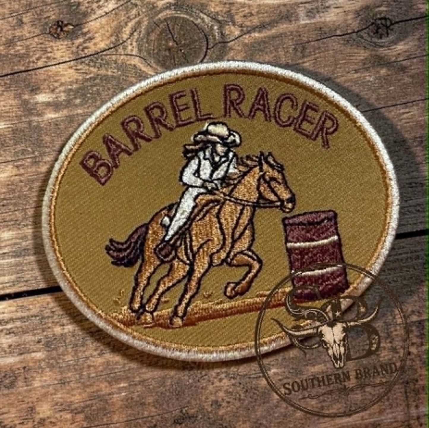 An oval embroidered patch featuring a cowboy riding a horse around a barrel, with the words "BARREL RACER" across the top. The patch is placed on a rustic wooden surface, with a circular "Southern Brand" watermark overlay in the lower right corner.