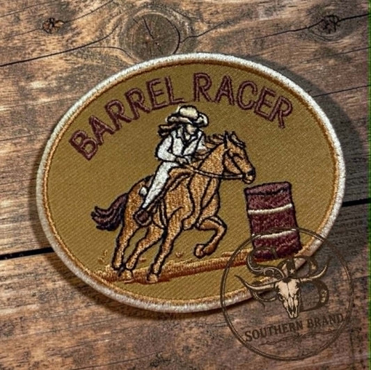 An oval embroidered patch featuring a cowboy riding a horse around a barrel, with the words "BARREL RACER" across the top. The patch is placed on a rustic wooden surface, with a circular "Southern Brand" watermark overlay in the lower right corner.