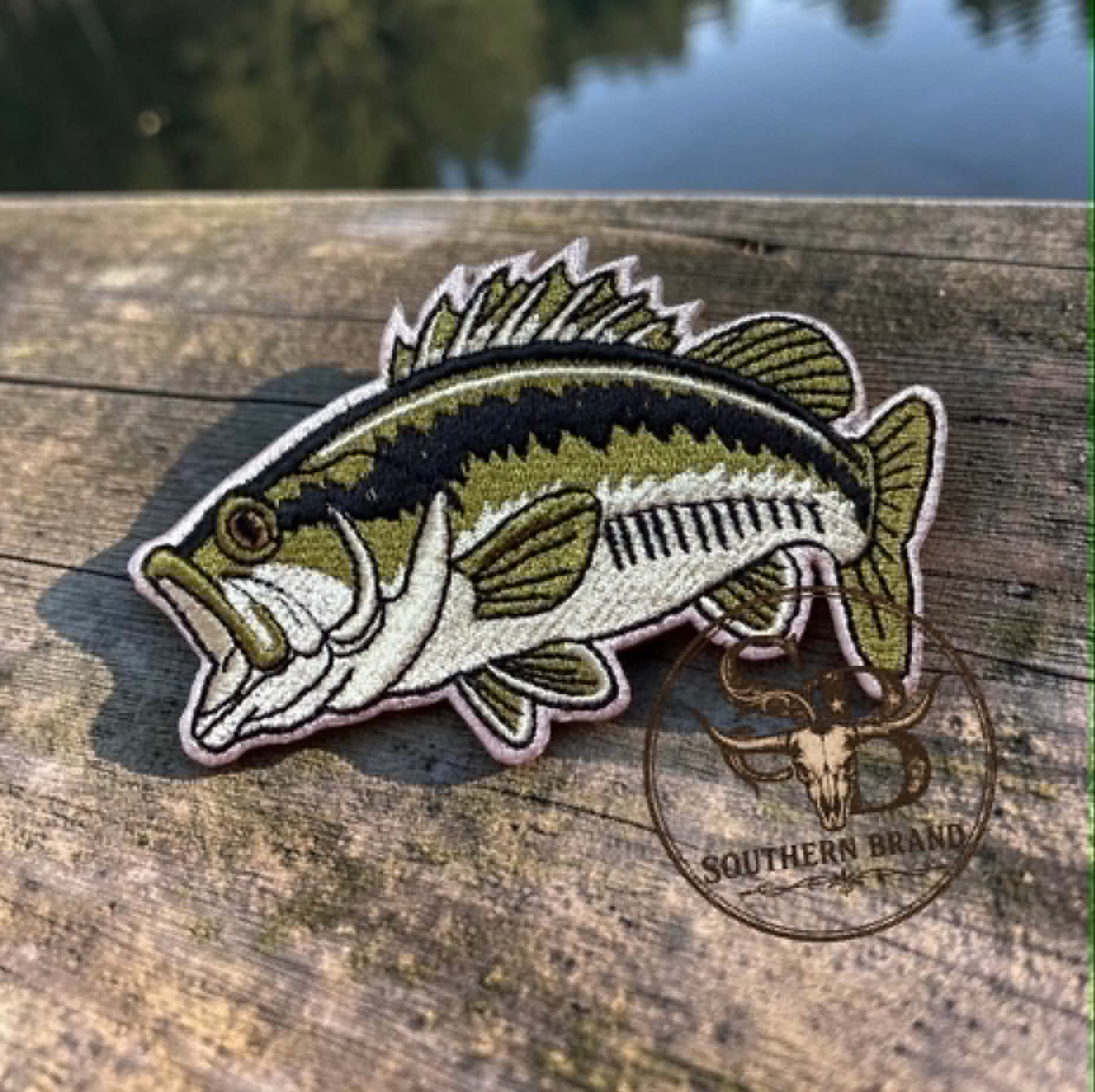 An embroidered bass fish patch with olive green, black, white, and tan details, resting on a weathered wooden dock with water and trees blurred in the background. A Southern Brand logo is visible on the patch.