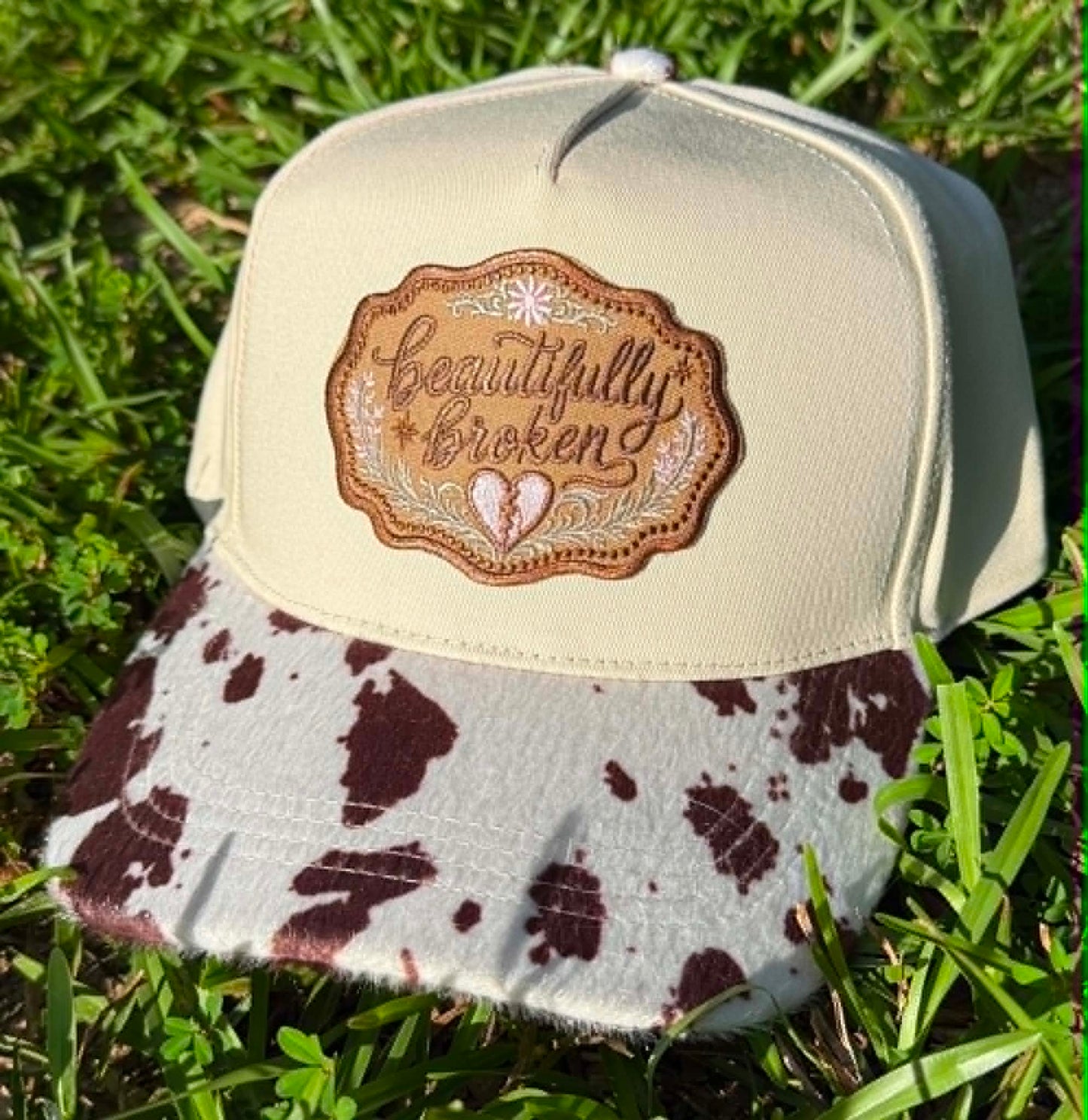 Cap with cow print brim and 'Beautifully Broken' patch on grass