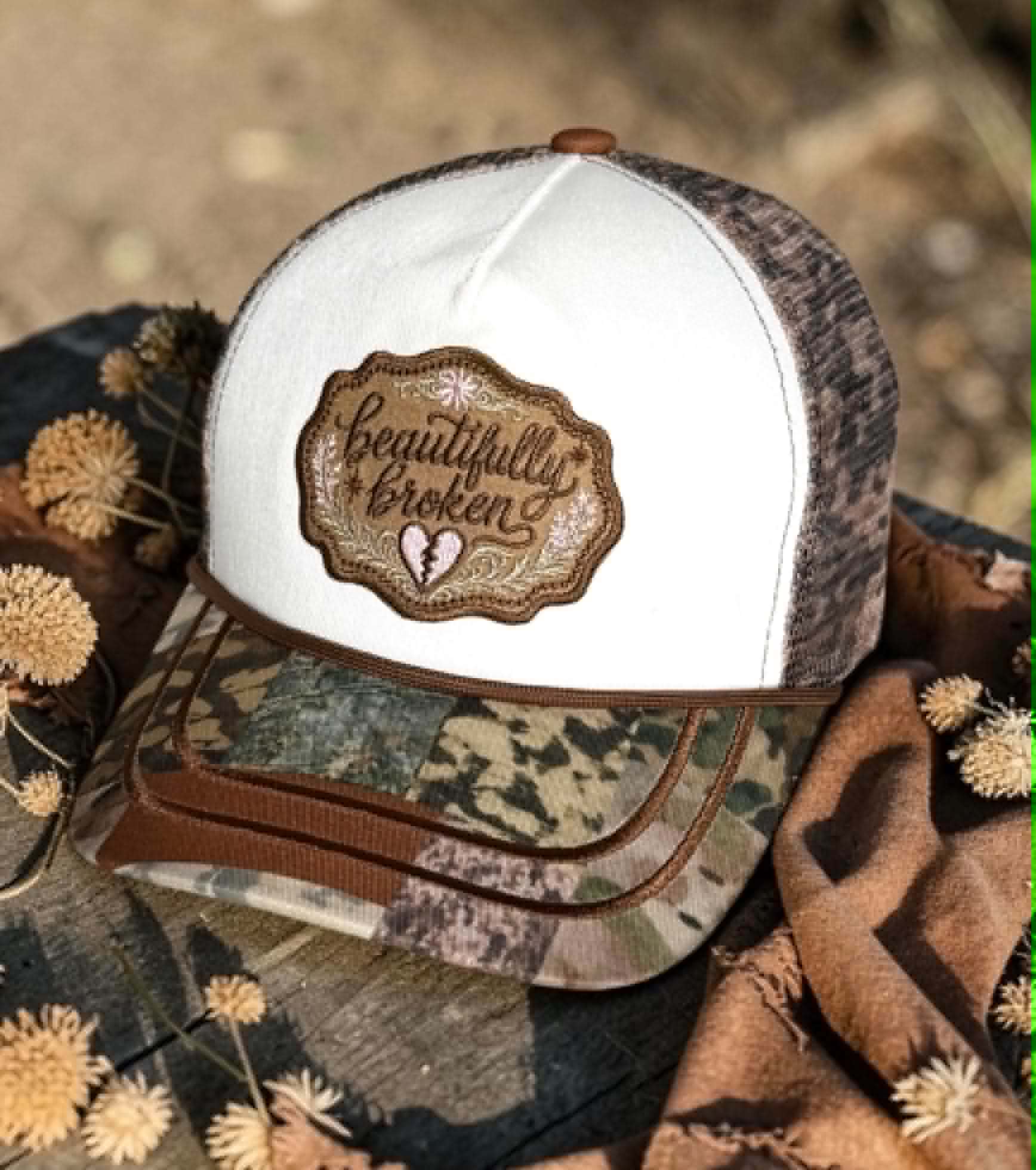 Camouflage and white cap with 'Beautifully Broken' design on a natural background