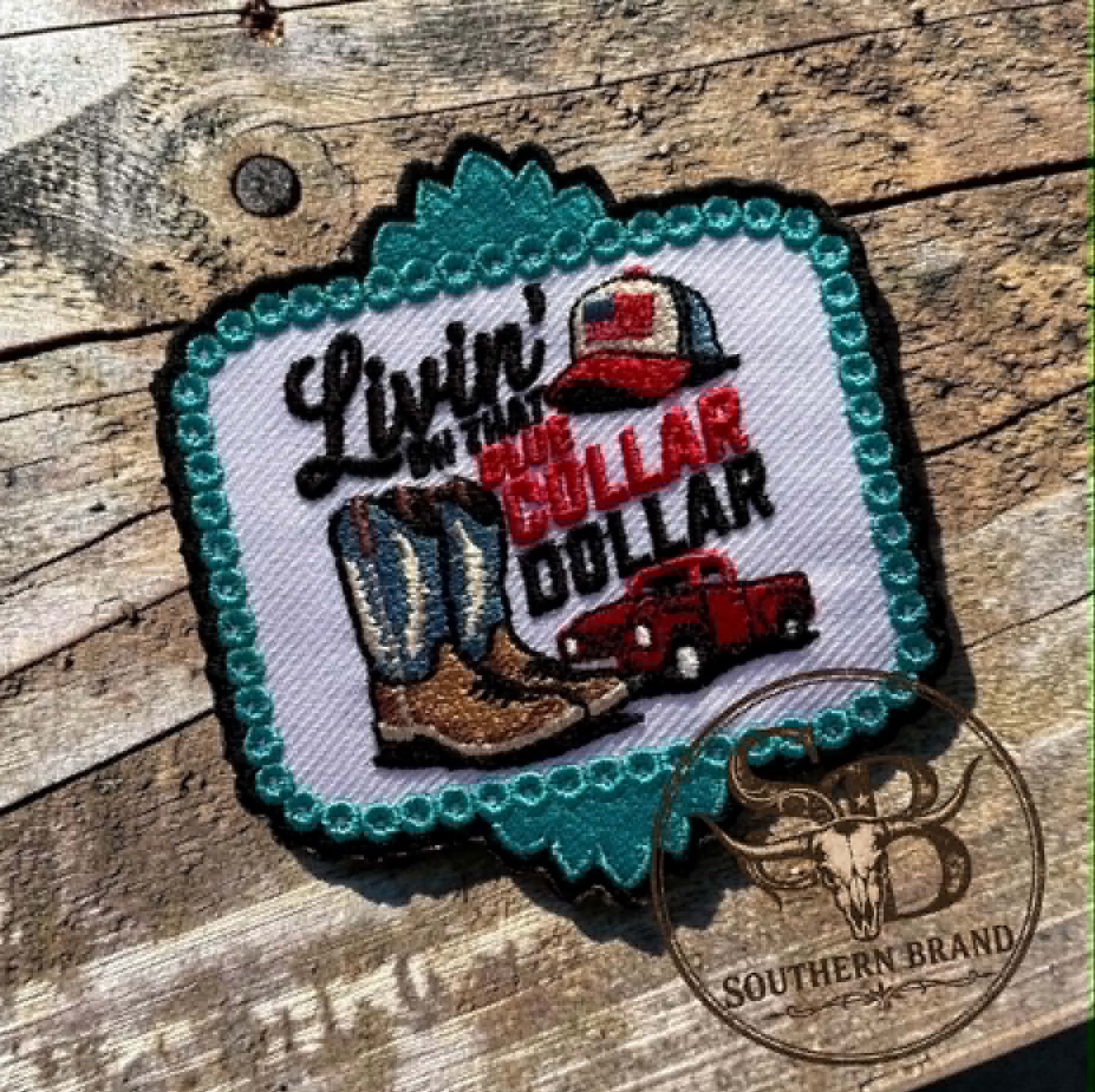 A decorative embroidered patch with a turquoise scalloped border and western-themed design reading "Livin' in the Bull Dollar," featuring cowboy boots, a red pickup truck, and a hat, placed on a rustic wooden surface.