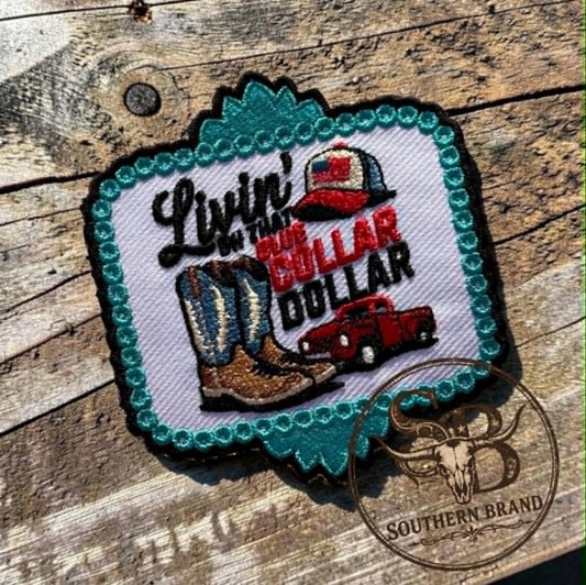 A decorative embroidered patch with a turquoise scalloped border and western-themed design reading "Livin' in the Bull Dollar," featuring cowboy boots, a red pickup truck, and a hat, placed on a rustic wooden surface.