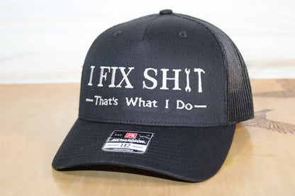Black cap with text 'I FIX SHIT That's What I Do' on a wooden surface