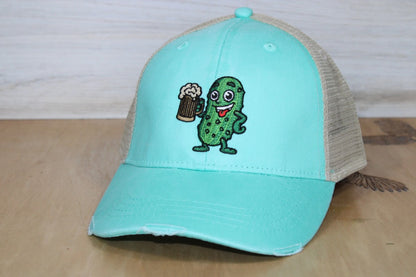 Turquoise cap with a pickle character holding a beer mug on a wooden surface.
