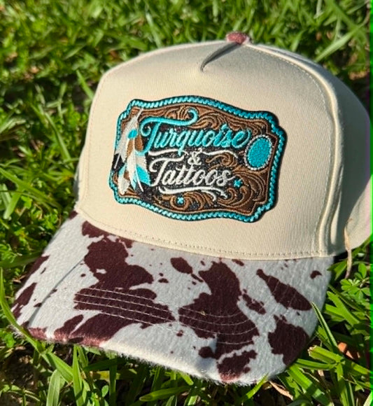 Beige cap with a cow print brim and 'Turquoise & Tattoos' logo on grass